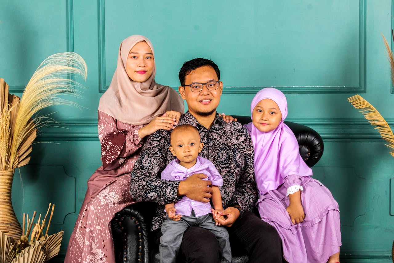 Smiling Muslim family of four posing indoors against a green wall.