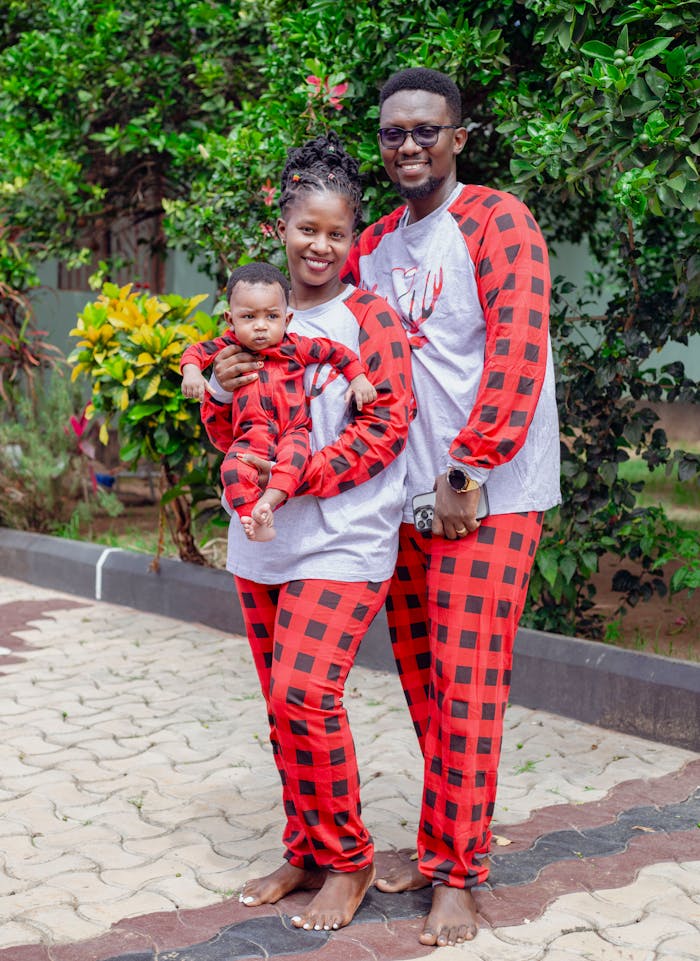 Family of three wearing red plaid pajamas posing outdoors with green foliage.